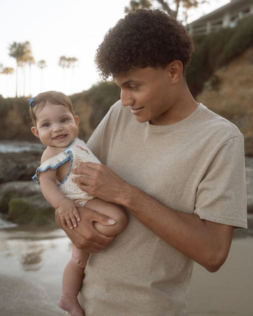 Teenage son smiling down at baby in his arms on beach with palm trees in background