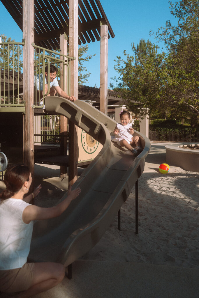 Family playground session, toddler on slide with parents
