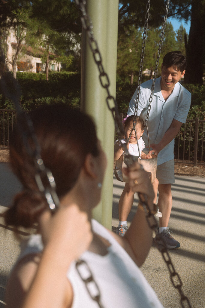 Dad pushing daughter on swing at neighborhood park with mother in foreground