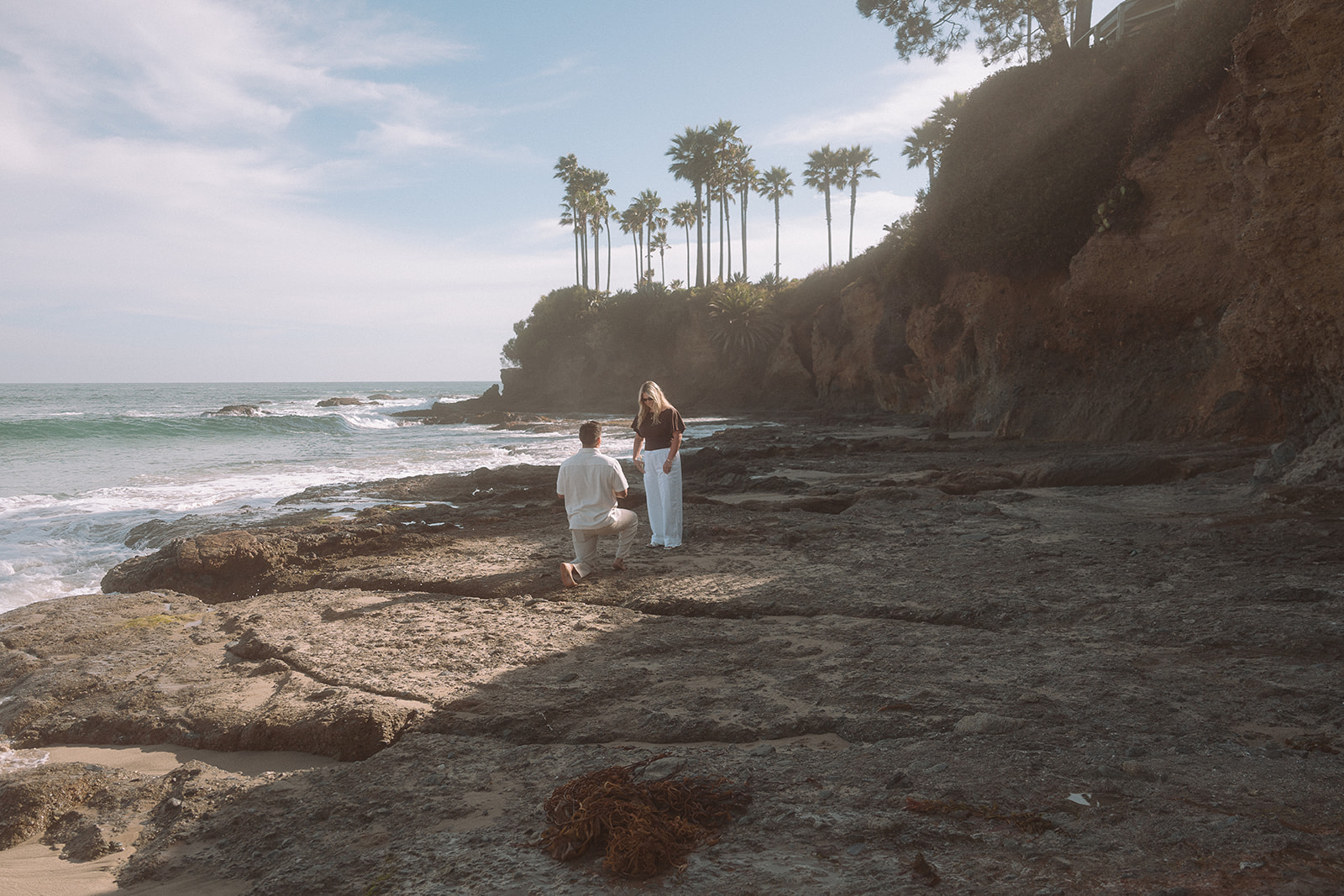 Man kneeling on the rocky shoreline proposes to his partner as a large wave crashes behind them at Shaws Cove in Laguna Beach.
