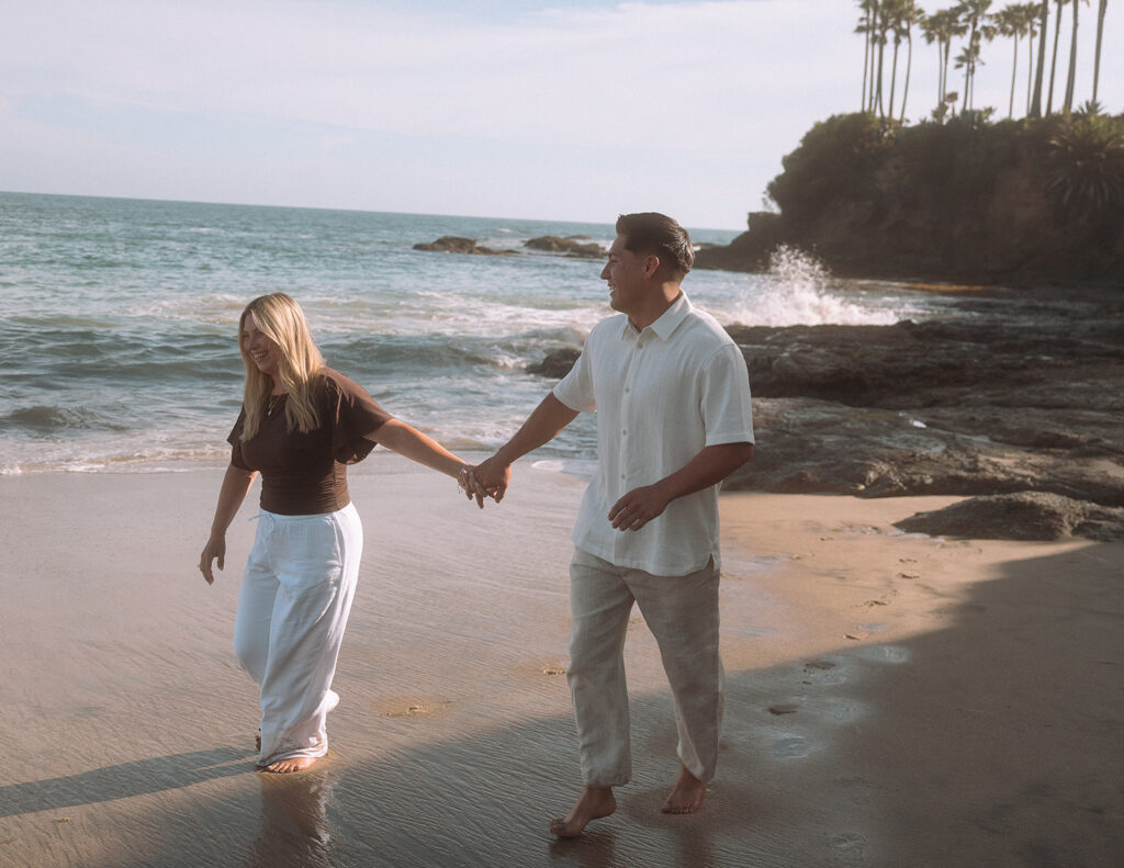 Newly engaged couple laughs while holding hands and walking along the shoreline at Shaws Cove, Laguna Beach.