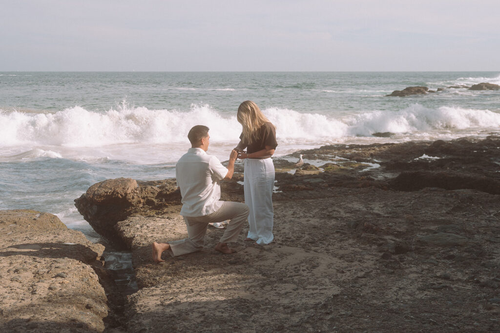 Man kneeling on rocky shore proposes to woman as a large wave crashes behind them at Shaws Cove in Laguna Beach.