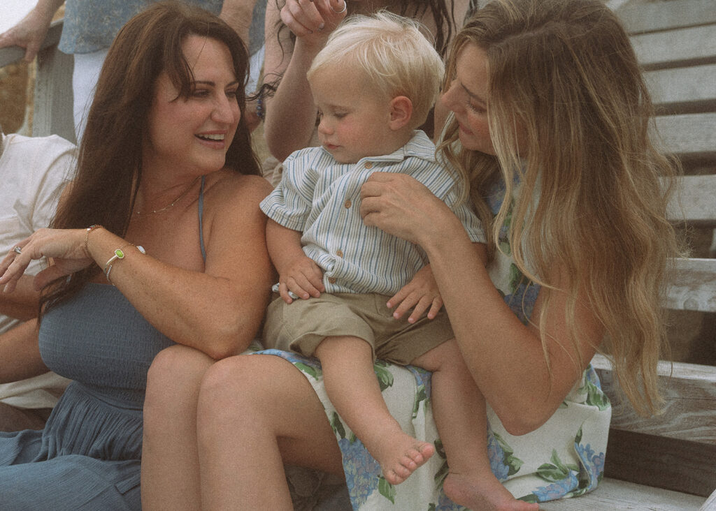 Three generations of women gathered around a toddler on the beach stairs, leaning in and smiling