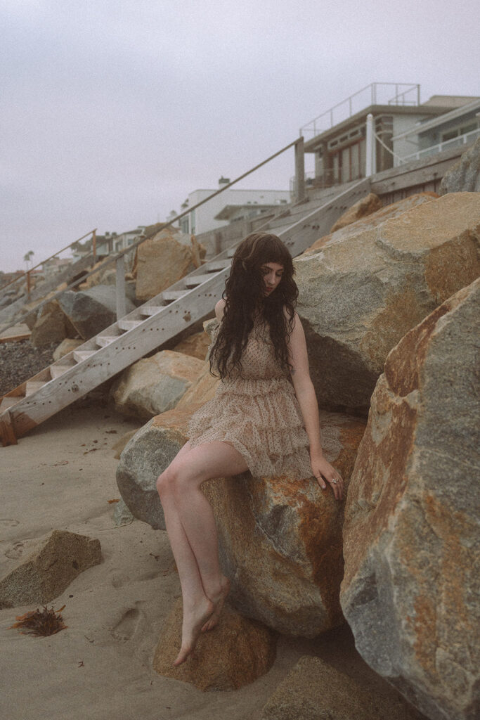 Woman in a soft tan dress resting against large granite boulders below the beach houses at Capistrano Beach