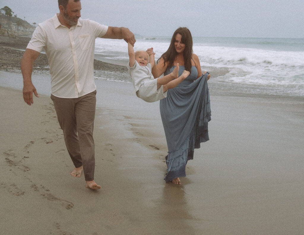 Grandparents swinging laughing toddler between them while walking barefoot along the wet sand