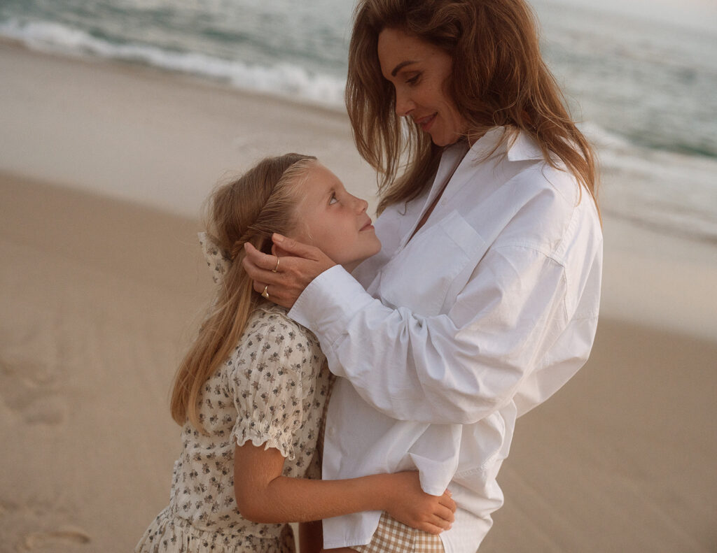 A mother holding her young daughter on the beach, the girl looking up at her while she smooths her hair.