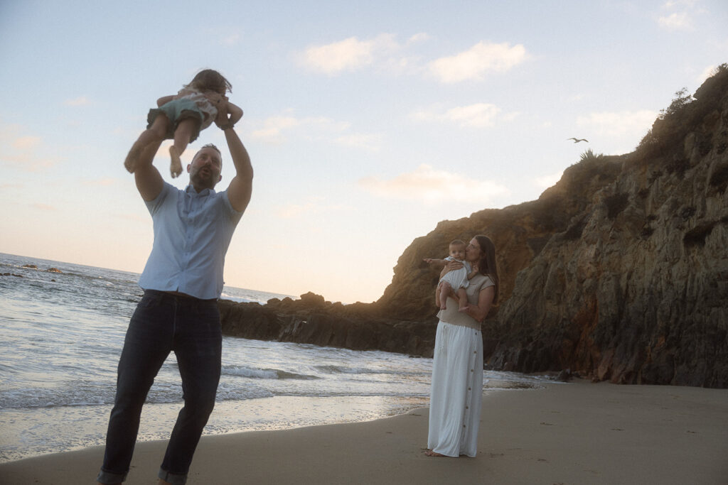 Family photos at Crescent Bay beach at sunset in Orange County. A dad in a light blue shirt and dark jeans lifts his toddler overhead, while a mom in a taupe knit top and white maxi skirt kisses the baby she's holding, with golden cliffs behind them.