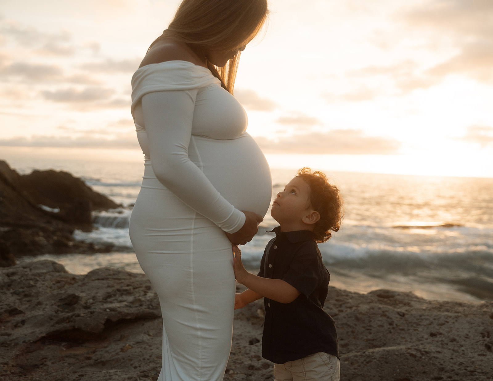 Toddler boy looking up at his pregnant mom with his hands on her belly during a second baby maternity session at Goff Cove in Laguna Beach.