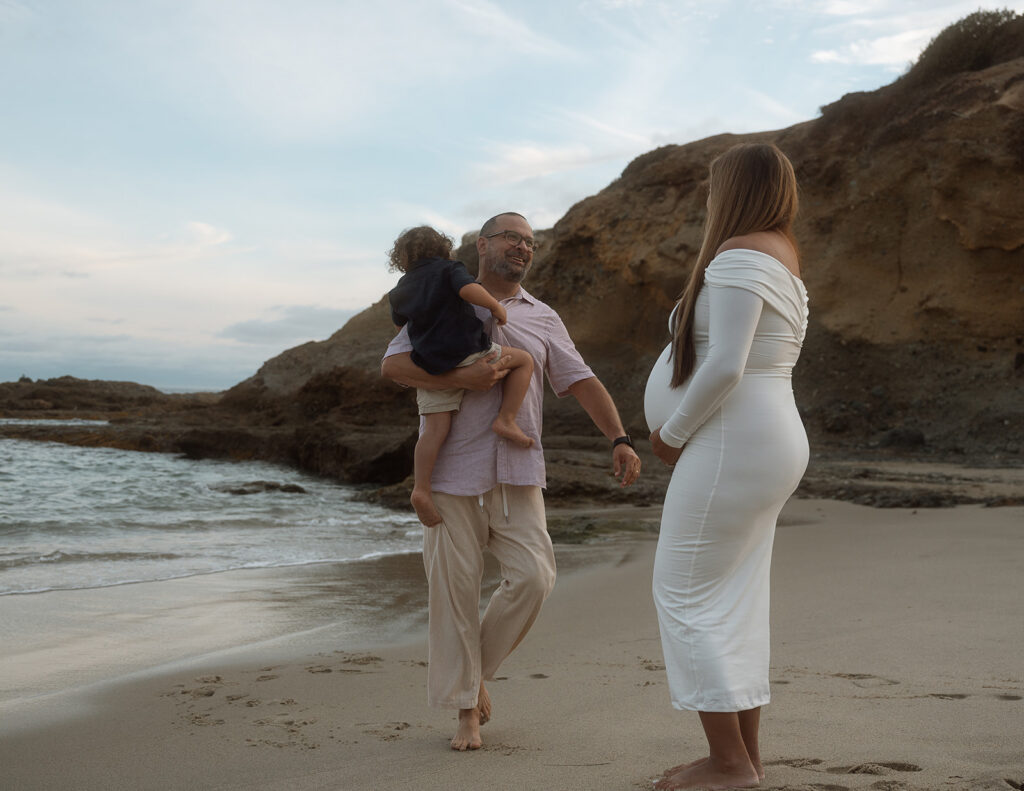 Pregnant mom watching her husband carry their toddler son along the sand during an Orange County maternity photo session.
