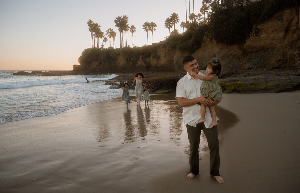 Dad carrying his toddler daughter along the shoreline while the rest of the family walks near the tide pools