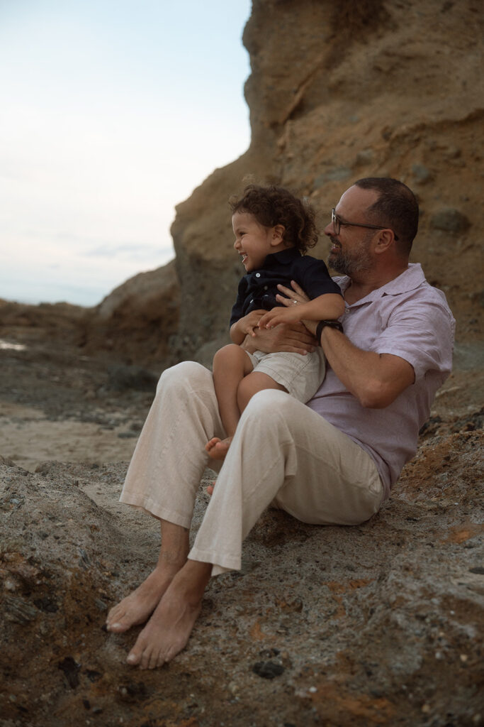 Dad sitting in the rocks holding his laughing toddler son during a beach family session in Laguna Beach.