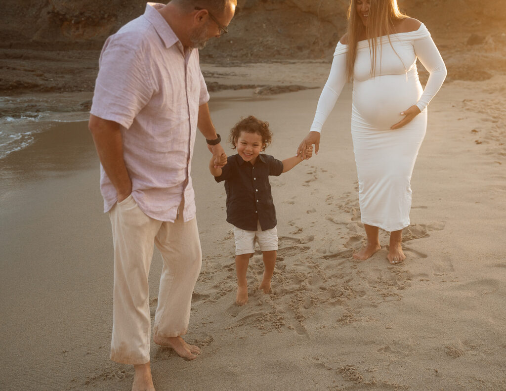 Expecting family of three walking barefoot along the shoreline below the cliffs during a golden hour maternity session in Laguna Beach.