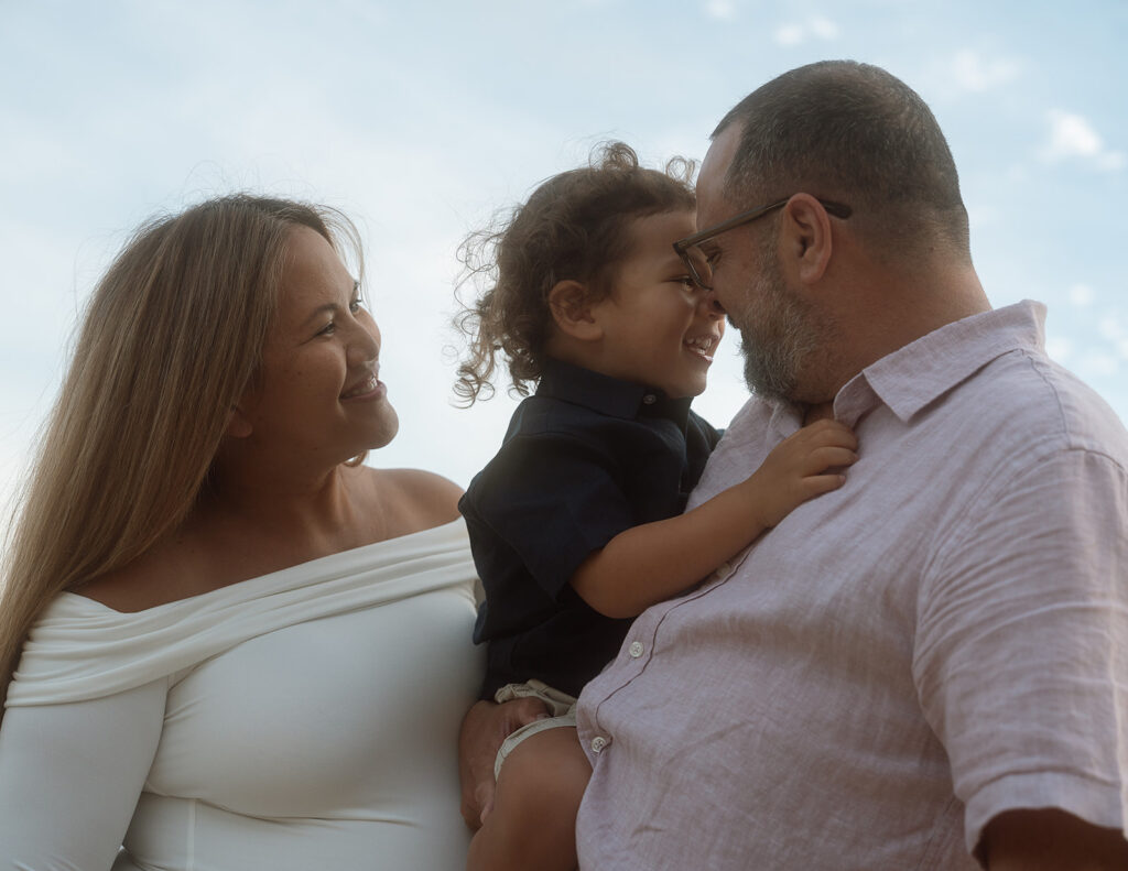Expecting parents laughing with their toddler son against an open sky during a family maternity session in Laguna Beach.