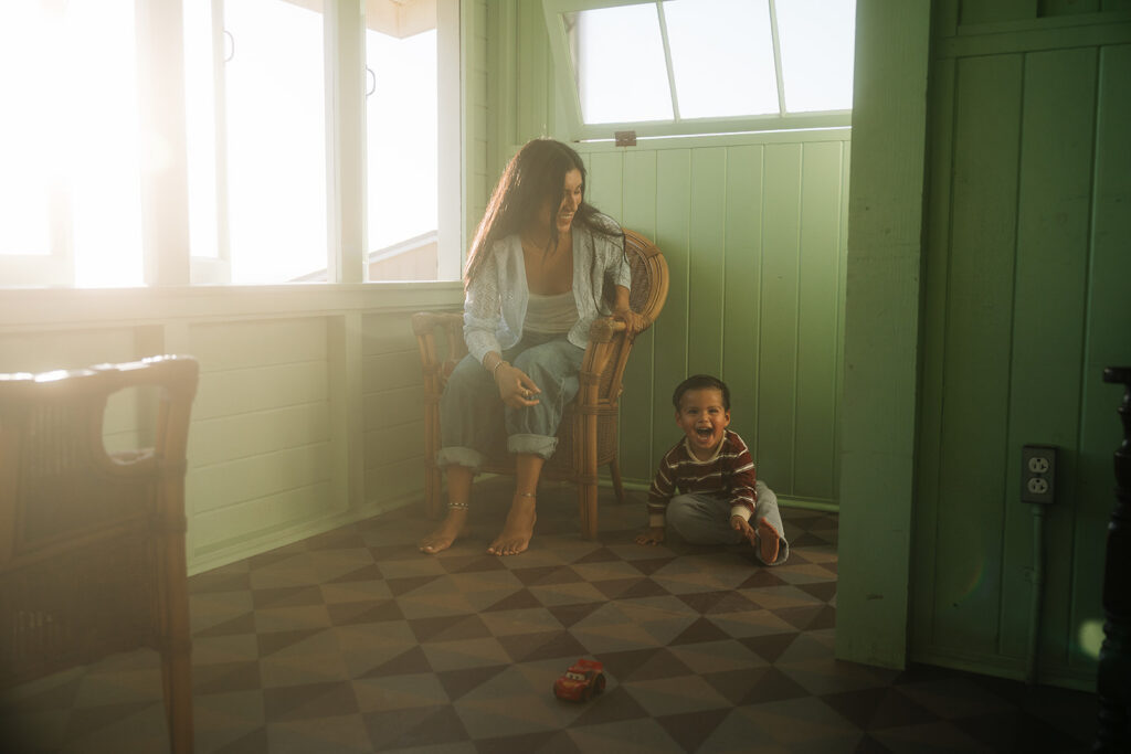Toddler laughing on the checkered floor while his mom smiles from a wicker chair in a sunlit Crystal Cove beach cottage, a lifestyle family photo in Newport Beach.