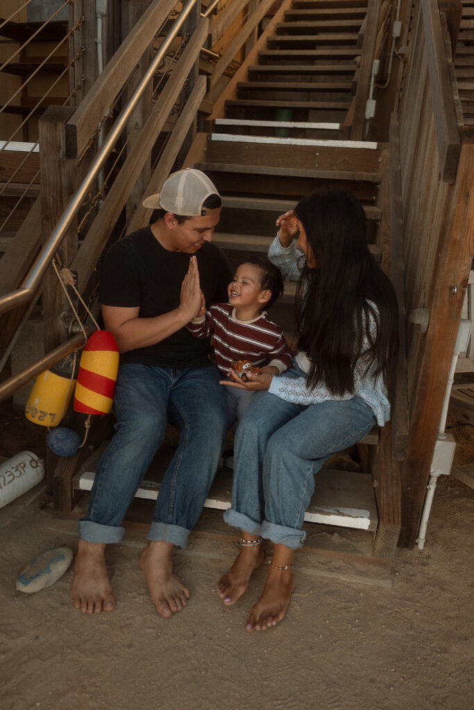 Dad high-fiving his laughing toddler on the wooden stairs of the Crystal Cove cottages, with mom beside them and colorful buoys hanging nearby.