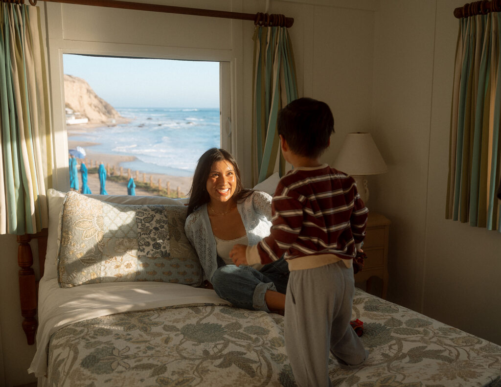 Mom grinning at her young son as he climbs onto the bed in a Crystal Cove cottage with an ocean view, a candid family photo in Orange County.