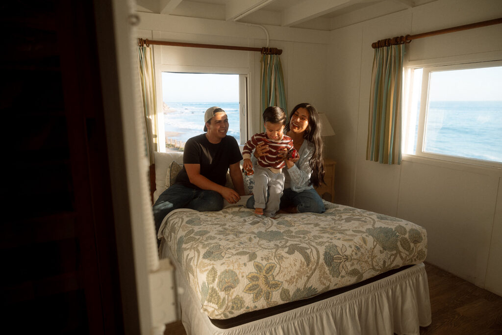 Parents laughing as their toddler stands on the bed in a Crystal Cove cottage bedroom with ocean views through both windows.