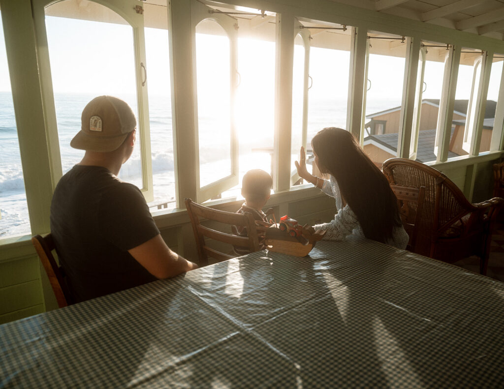 Family sitting at the dining table watching the waves through open cottage windows at Crystal Cove State Park.