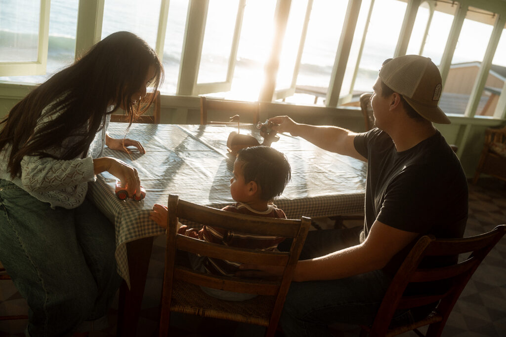 Mom and dad playing toy cars with their toddler at the dining table of a sunlit Crystal Cove cottage, a candid family session in Newport Beach.