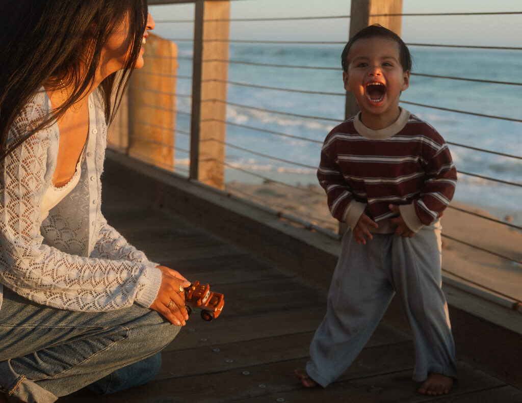 Barefoot toddler laughing with his mouth wide open on the ocean-view boardwalk at Crystal Cove, a candid family photo near Laguna Beach.