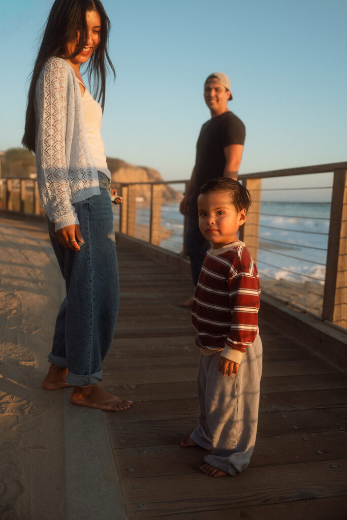 Toddler glancing back at the camera on the Crystal Cove boardwalk at golden hour while his parents smile behind him.