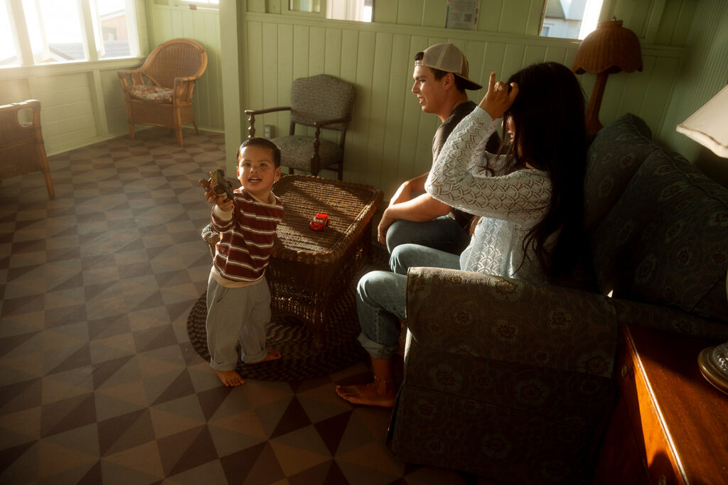 Toddler proudly holding up a toy car while his parents relax on the couch in a mint green beach cottage sunroom in Orange County.